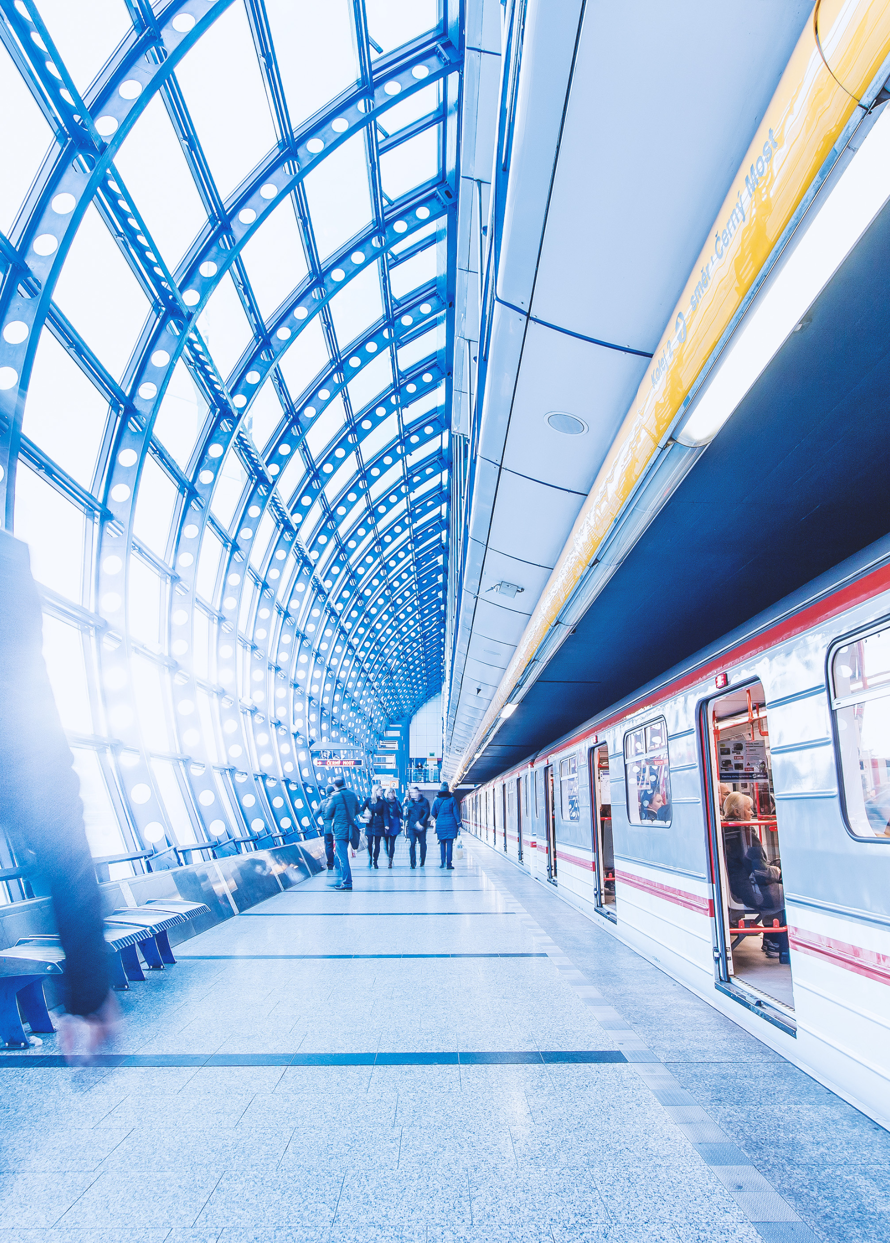 A modern and bright subway station with a glass and steel vaulted structure. A train is parked on the left track with some people seated inside, while others walk on the platform. The lighting is bright and the design is futuristic.