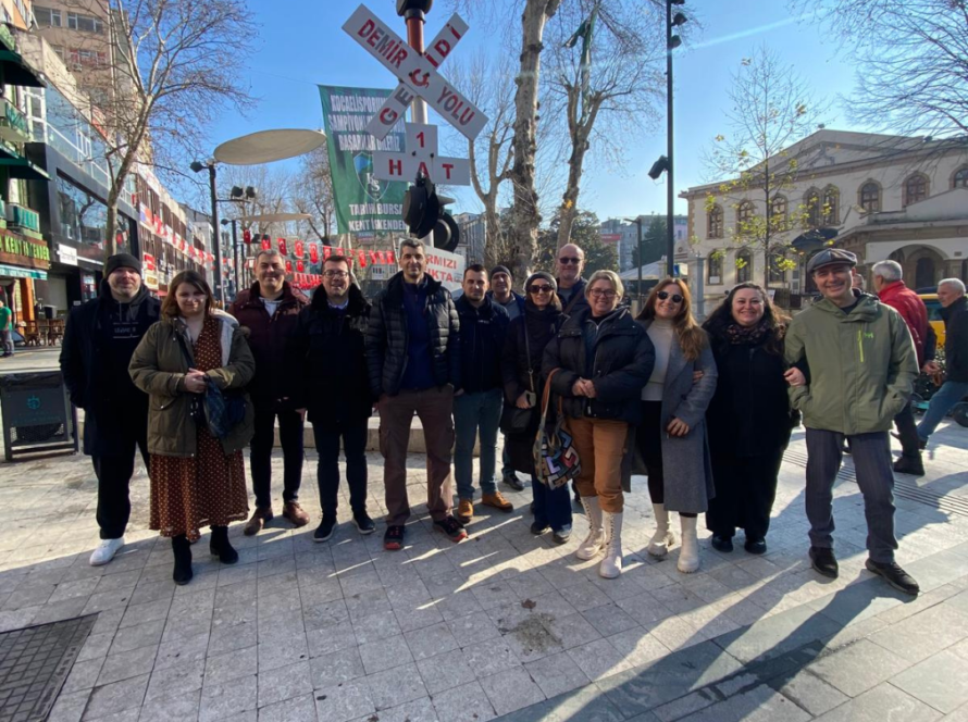 Partners of RISS project smiling in an urban area with a "Demir ve Yolu" sign and trees in the background